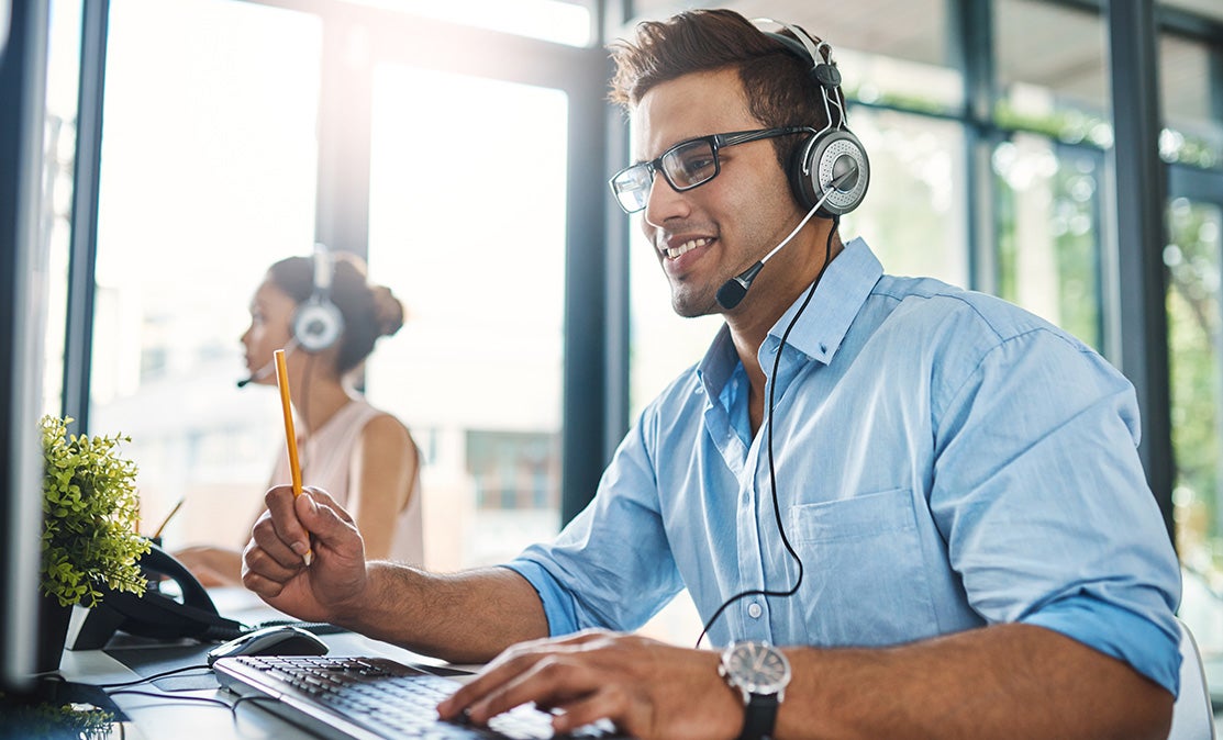 Man with headset smiling while working on a computer in a modern office. 